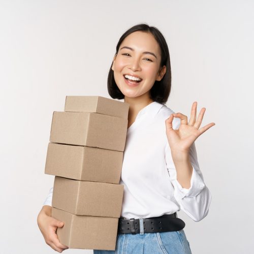 Portrait of beautiful happy woman carry boxes with orders. Businesswoman with carry delivery and showing okay sign, white background.